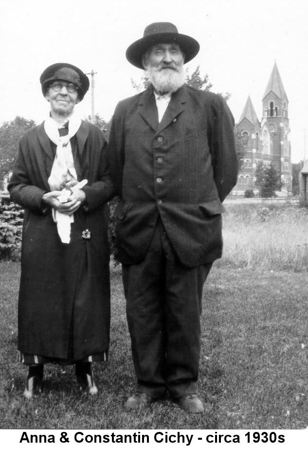 Black and white photo of an elderly woman and man standing together with a stone church in the distant background. The woman wears a black hat, large black-framed glasses, and an ankle-length dark loose-fitting dress with a large white shawl tied loosely around her neck and hanging to below her waist. Her hands are clasped at her waist and she has a close-lipped smile. The man has a very bushy white beard and moustace; he wears a baggy dark suit and a dark broad-brimmed hat; his hands are clasped behind his back and there is a grin on his lips and a twinkle in his eyes.